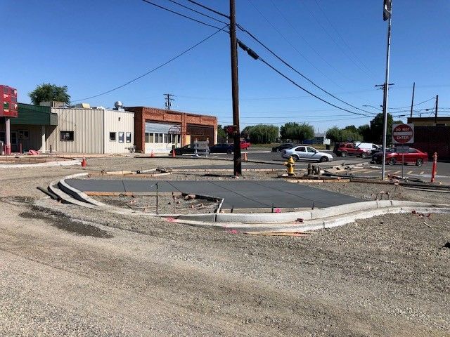 Concrete construction site at a street intersection, new sidewalk or median being formed, power lines and parked cars visible.