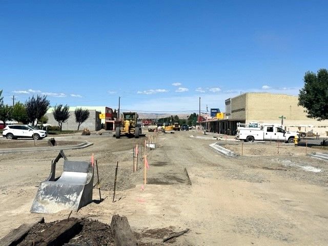Construction site on a city street. Heavy machinery, work trucks, and piles of gravel are visible. Blue sky overhead.