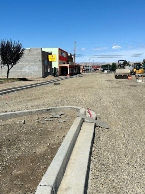 Road construction with gravel surface, concrete curb, buildings in background under blue sky.