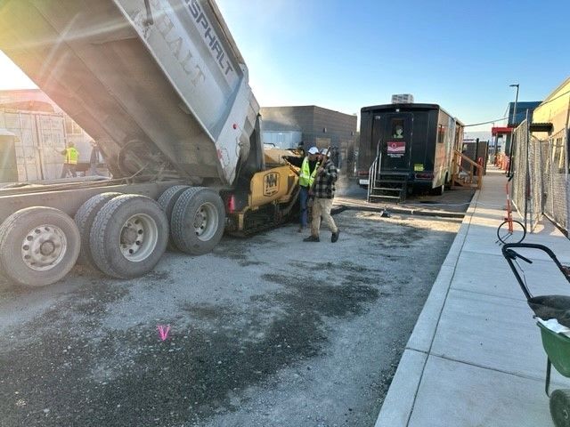 Truck dumping asphalt onto a construction site with workers nearby; a trailer is visible.