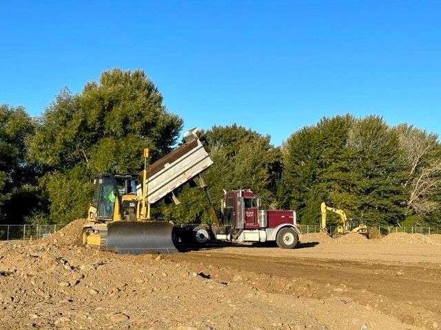 Bulldozer pushing dirt onto a dump truck; excavator in background, clear sky.