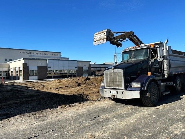 Dump truck and excavator near a school building under construction on a sunny day.