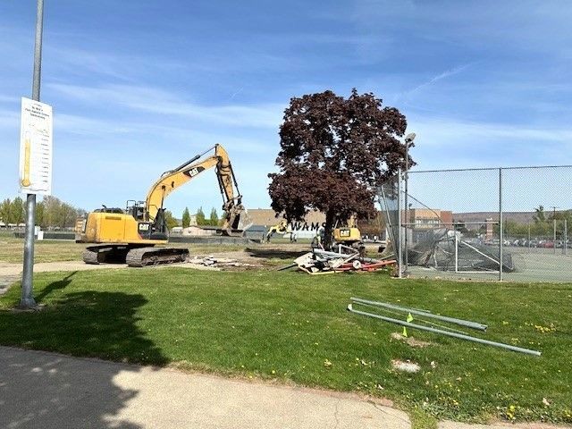 Excavator demolishing near a red-leaved tree, chain link fence, and green grass field on a sunny day.