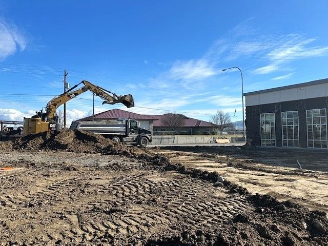 Construction site with excavator and dump truck; new building next to older one, sunny day.
