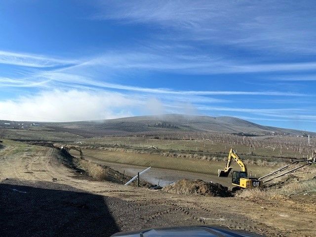 Construction site with excavator near a body of water, hill in background with cloudy sky.