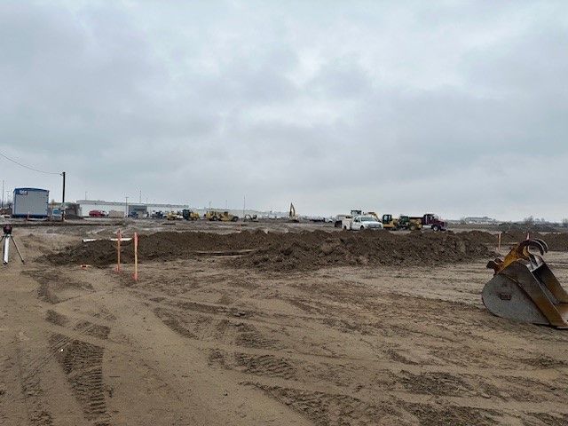Construction site under a cloudy sky with dirt mounds, heavy machinery, and distant buildings.