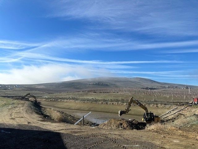 Construction site: Excavator digging near a water feature, with a hill and cloudy sky in the background.