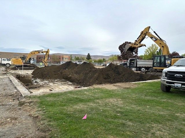 Construction site with excavators moving dirt into a dump truck, and a white pickup truck on grass.