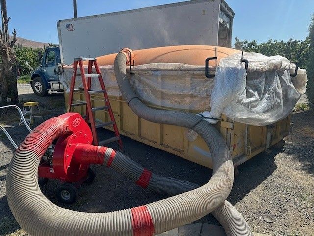 A large industrial container with a blower connected to a hose, likely for processing materials outdoors.