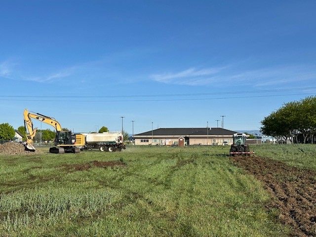 Construction site with excavator and tractor plowing field; building in background.