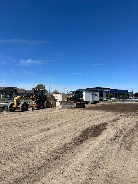 Construction site with a grader and bulldozer on dirt, with buildings and blue sky background.