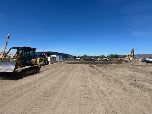 Construction site with bulldozer and excavator under a blue sky.  Graded dirt field with buildings in the background.
