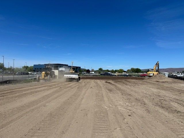 Construction site with dirt ground, heavy machinery, and a building under a blue sky.