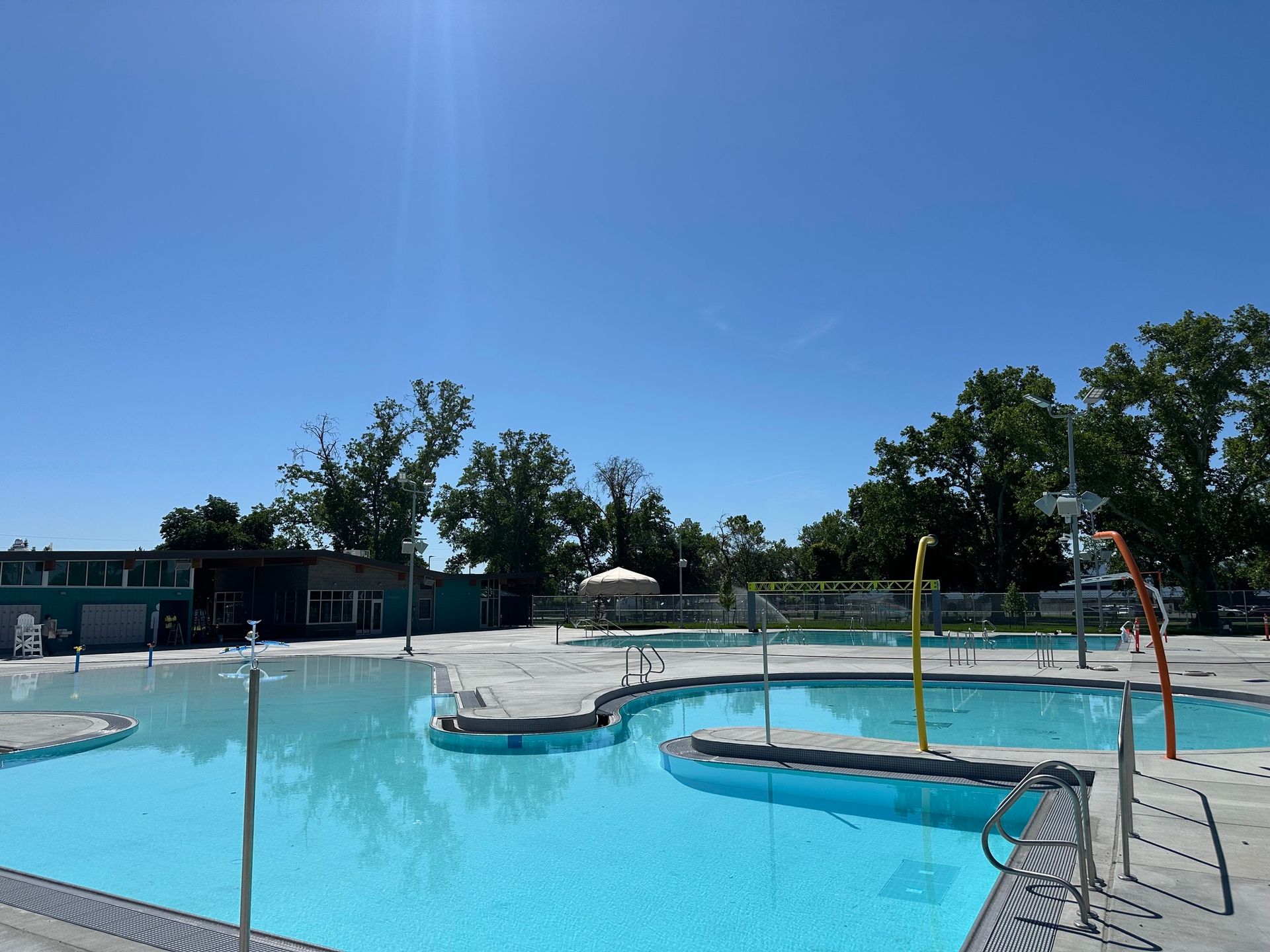 A public swimming pool on a sunny day. Light blue water, concrete deck, trees, and blue sky.