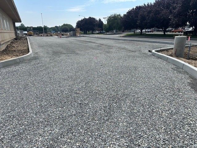 Gravel parking lot next to a building. Concrete curbs define edges. Trees and sky in the background.