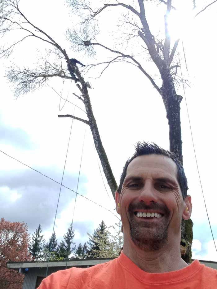 Man smiling in front of a tall tree; an arborist is pruning the tree in the background.