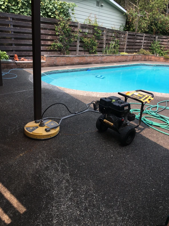 A pressure washer on a patio by a pool, connected to a yellow base, with a wooden fence in the background.