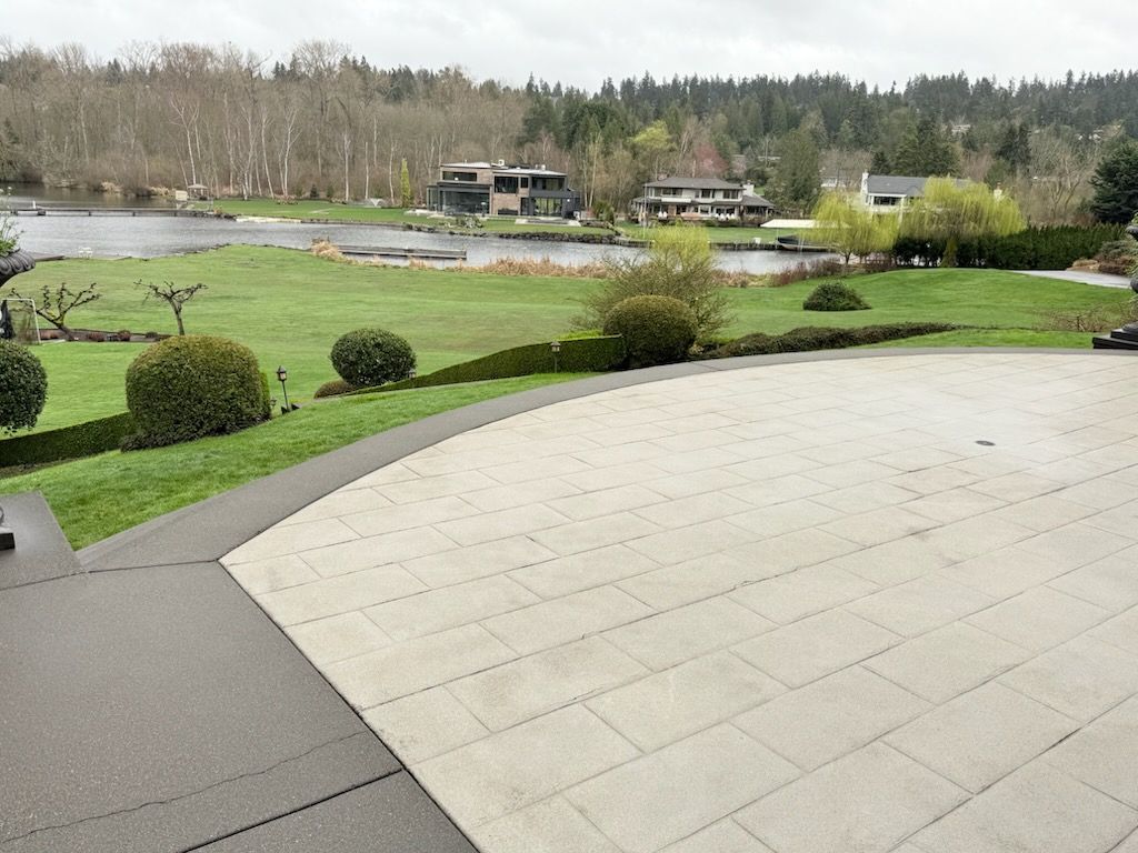 Overlooking a lake, a patio with white tiles and a green lawn with small shrubs; trees in the background.