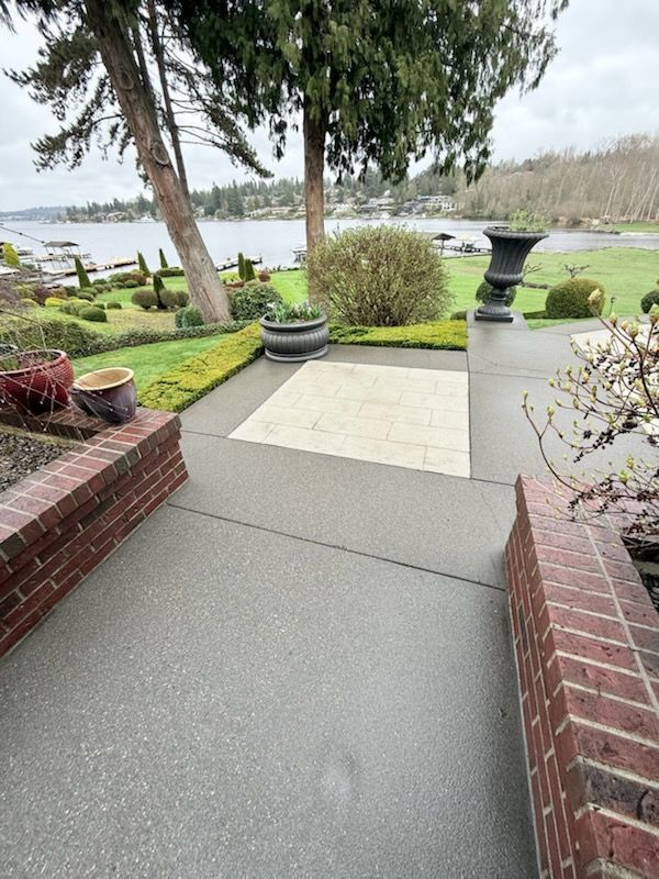 Exterior view of a paved walkway leading to a waterfront garden. Brick borders and trees frame the pathway.