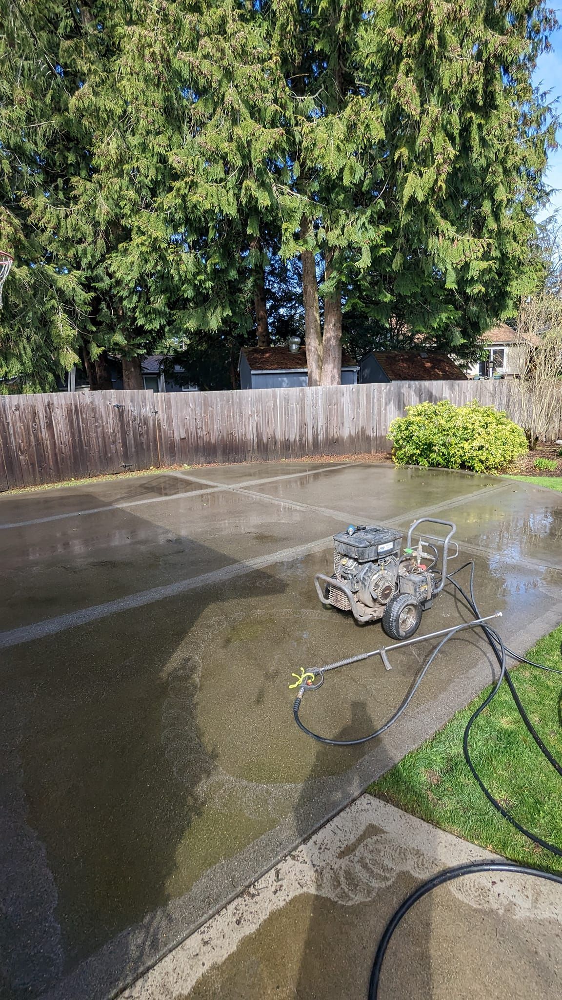 Man power washing a concrete patio, surrounded by a wooden fence, grass, and trees.