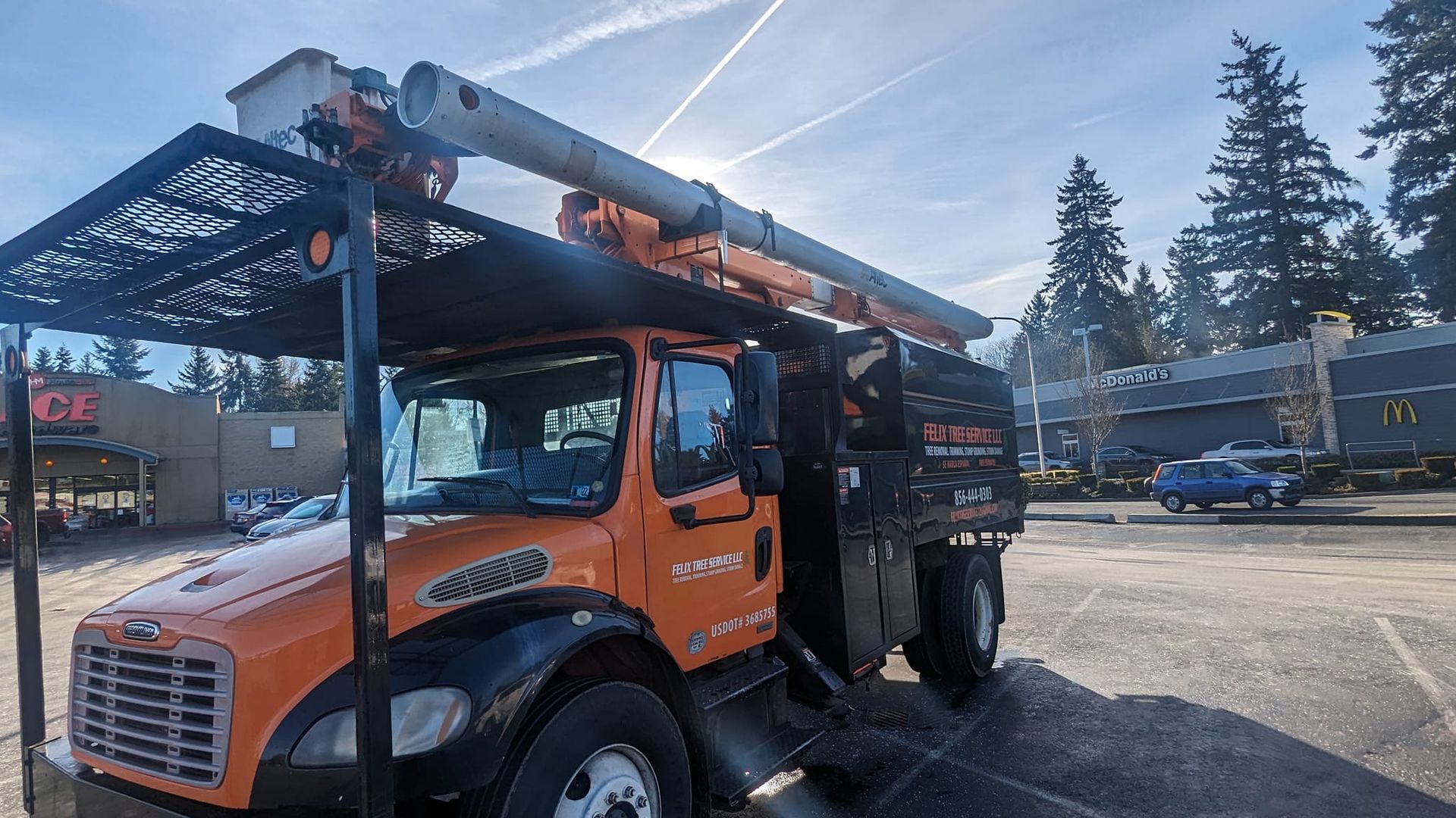 Orange tree service truck parked in a lot with a raised arm. Bright sunlight.