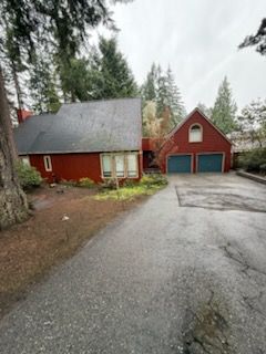 Red house and detached garage on a paved driveway, surrounded by trees.