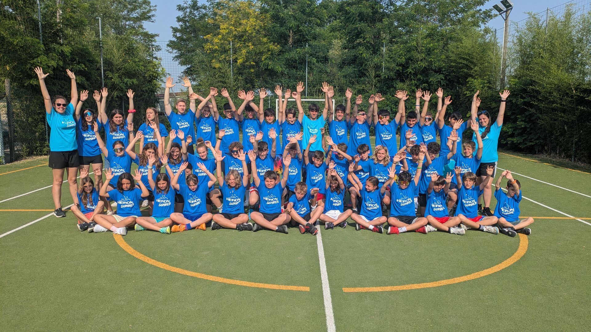 Un folto gruppo di ragazzi su una spiaggia alzano le braccia sorridendo. Cielo coperto, sabbia e mare sullo sfondo.