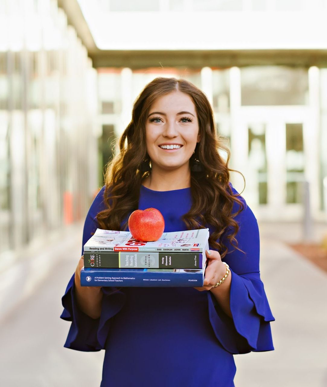 A woman in a blue dress is holding a stack of books and an apple.