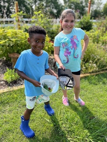 Montessori children are standing in the grass holding vegetables.