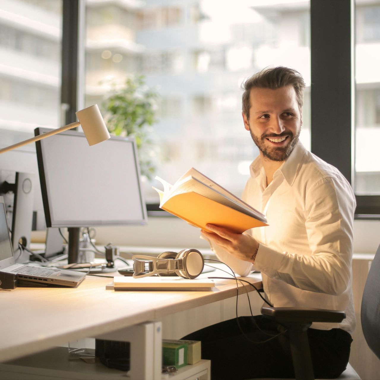 A man is sitting at a desk holding a book and smiling.