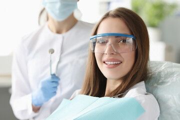 Woman smiles in a dental chair, wearing safety glasses. A dentist in a mask and gloves stands behind her, holding dental tools.
