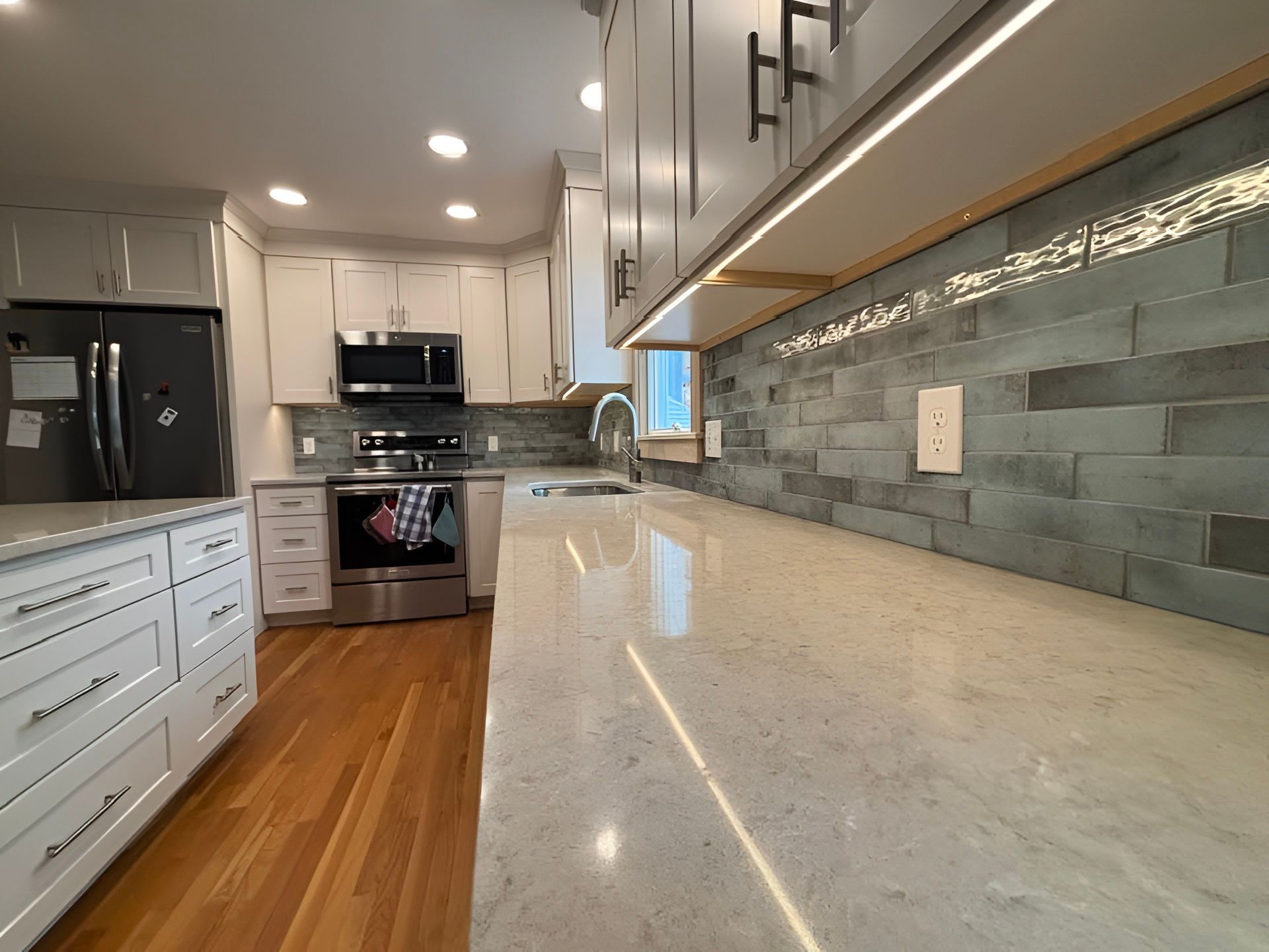 Bright modern kitchen with white cabinets, stainless appliances, wood floor, and long stone countertop