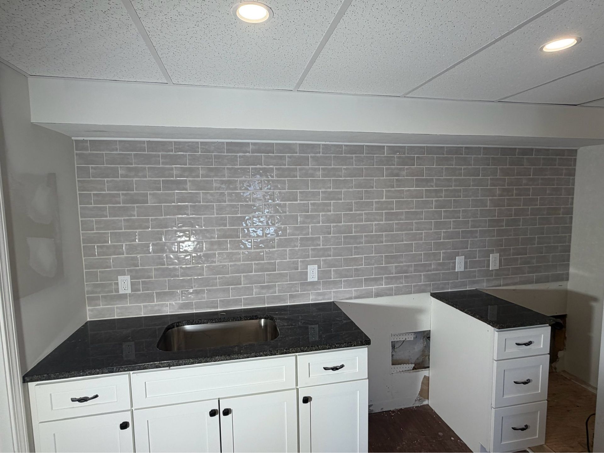 Empty kitchen with white cabinets, black countertops, gray subway tile backsplash, and a sink cutout at center