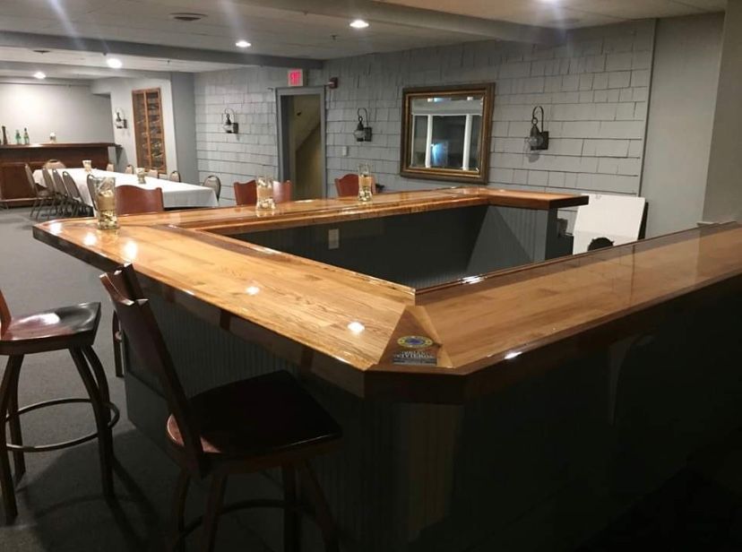 Empty bar interior with a wooden U-shaped counter and stools, lit by ceiling lights and wall sconces.