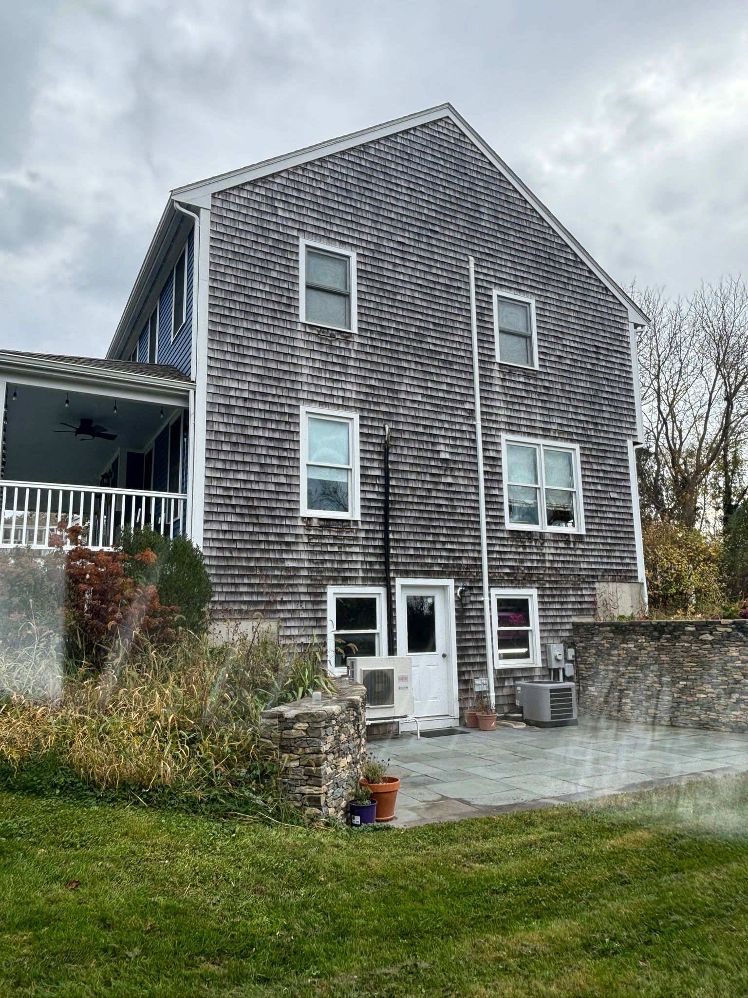 Tall gray-shingled house with white-trim windows, porch, and gravel yard under cloudy sky