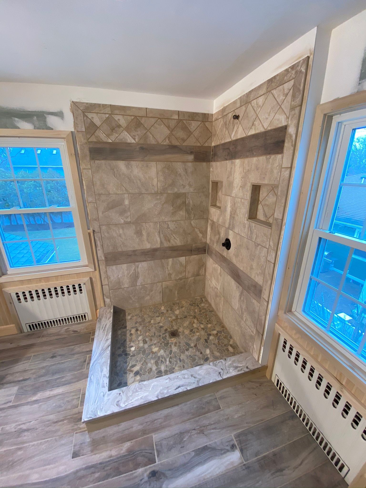 Stone-tiled walk-in shower in a bathroom with wood flooring and windows on both sides.