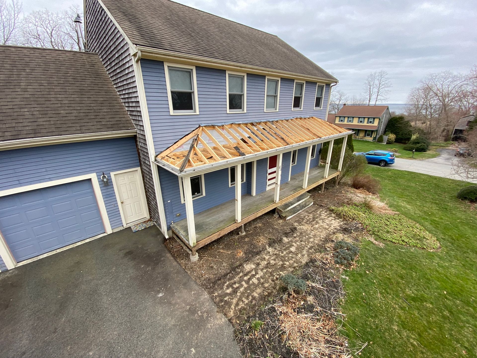 Blue two-story house with a damaged front porch roof, garage, and sloped yard under cloudy skies