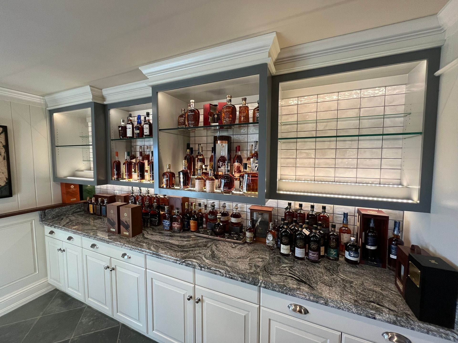 Home bar with white cabinets, granite countertop, illuminated shelves of liquor bottles, and framed decor.