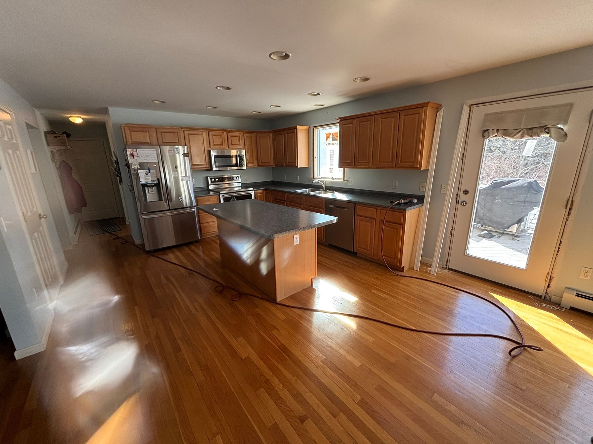 Empty kitchen with hardwood floors, wood cabinets, island, and large glass door to a snowy deck.