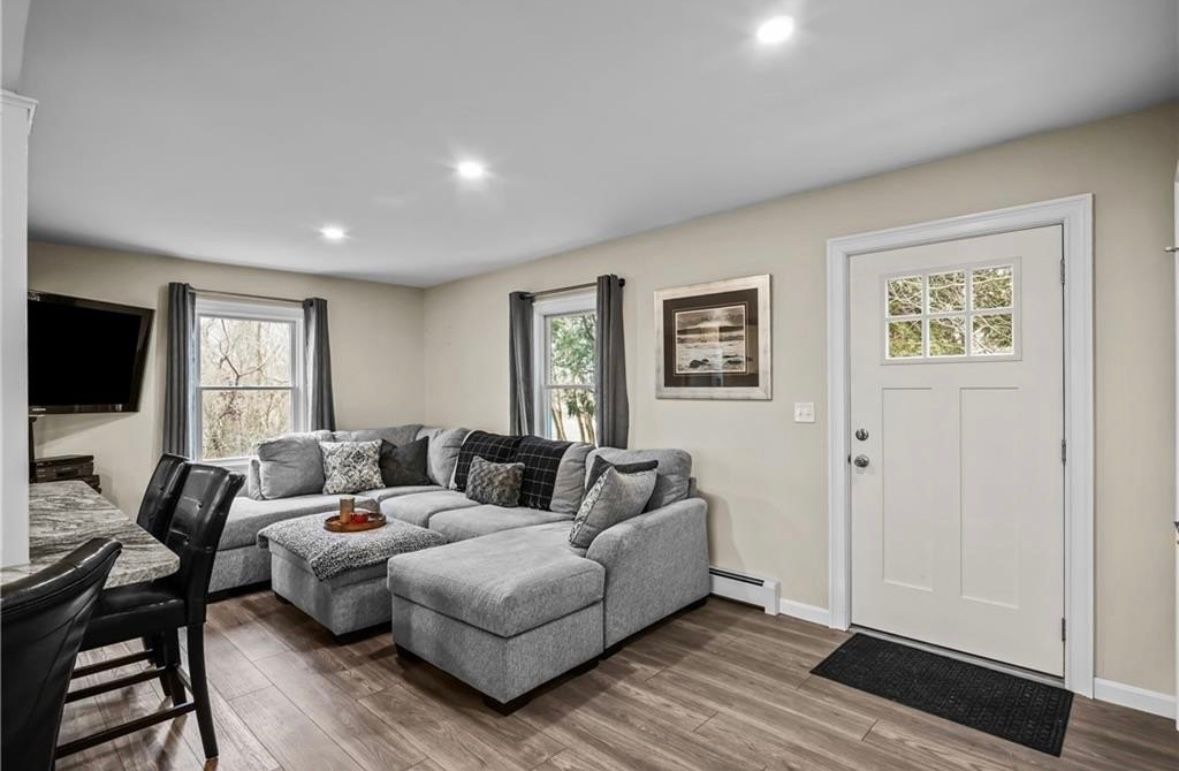 Bright living room with gray sectional sofa, wall-mounted TV, windows, and white front door