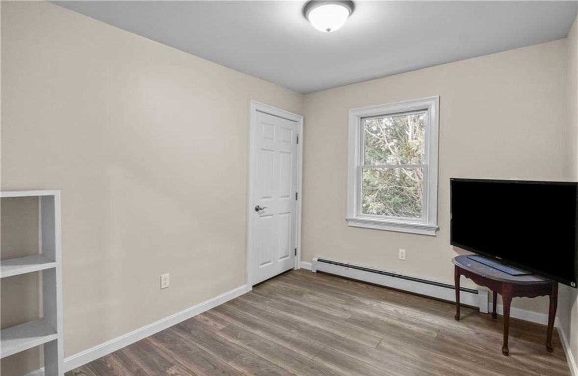 Empty beige bedroom with white door, window, TV on a small table, and wood-look floor.