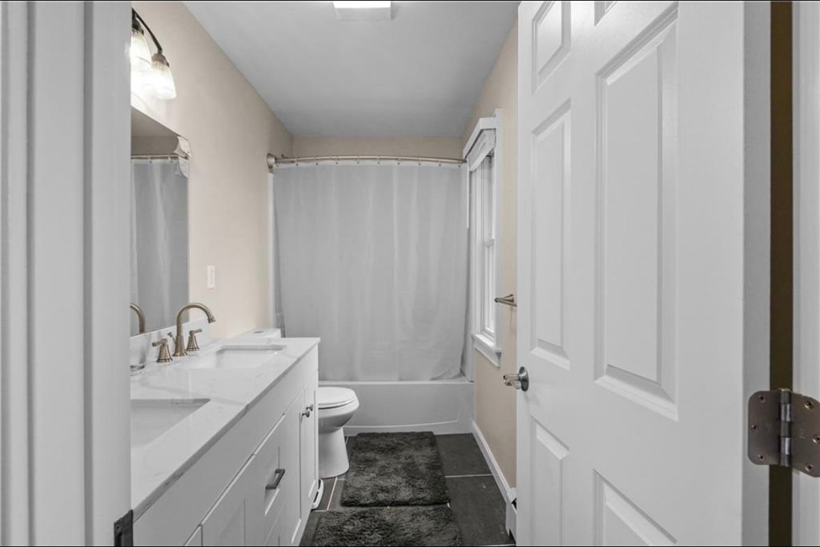 Narrow bathroom with white vanity, toilet, and shower curtain on a dark rug.