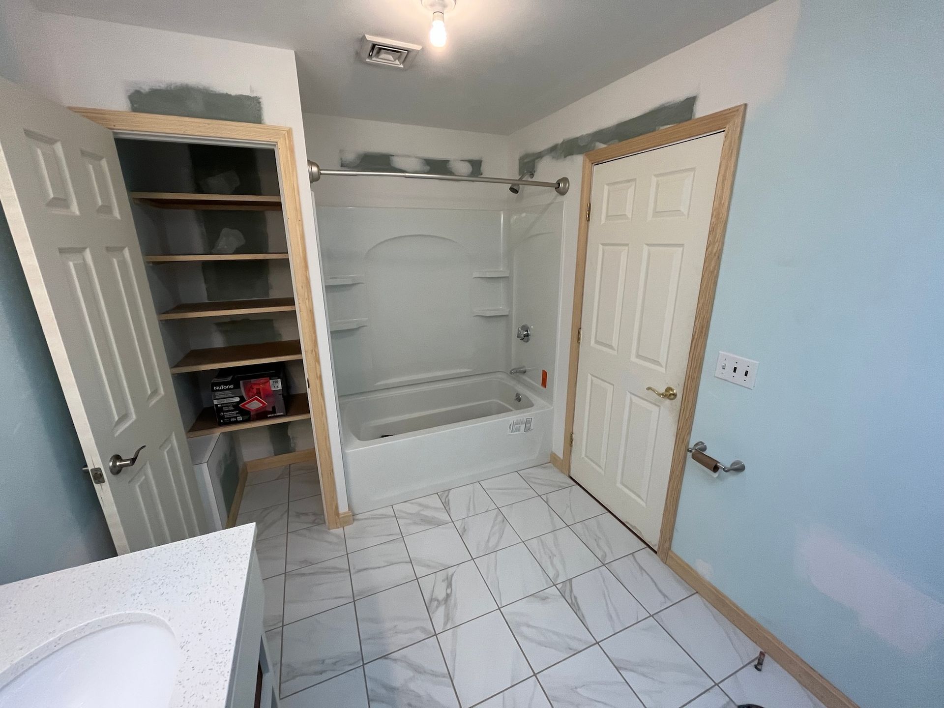 Small bathroom with tiled floor, white tub-shower, open linen closet, and a light blue wall