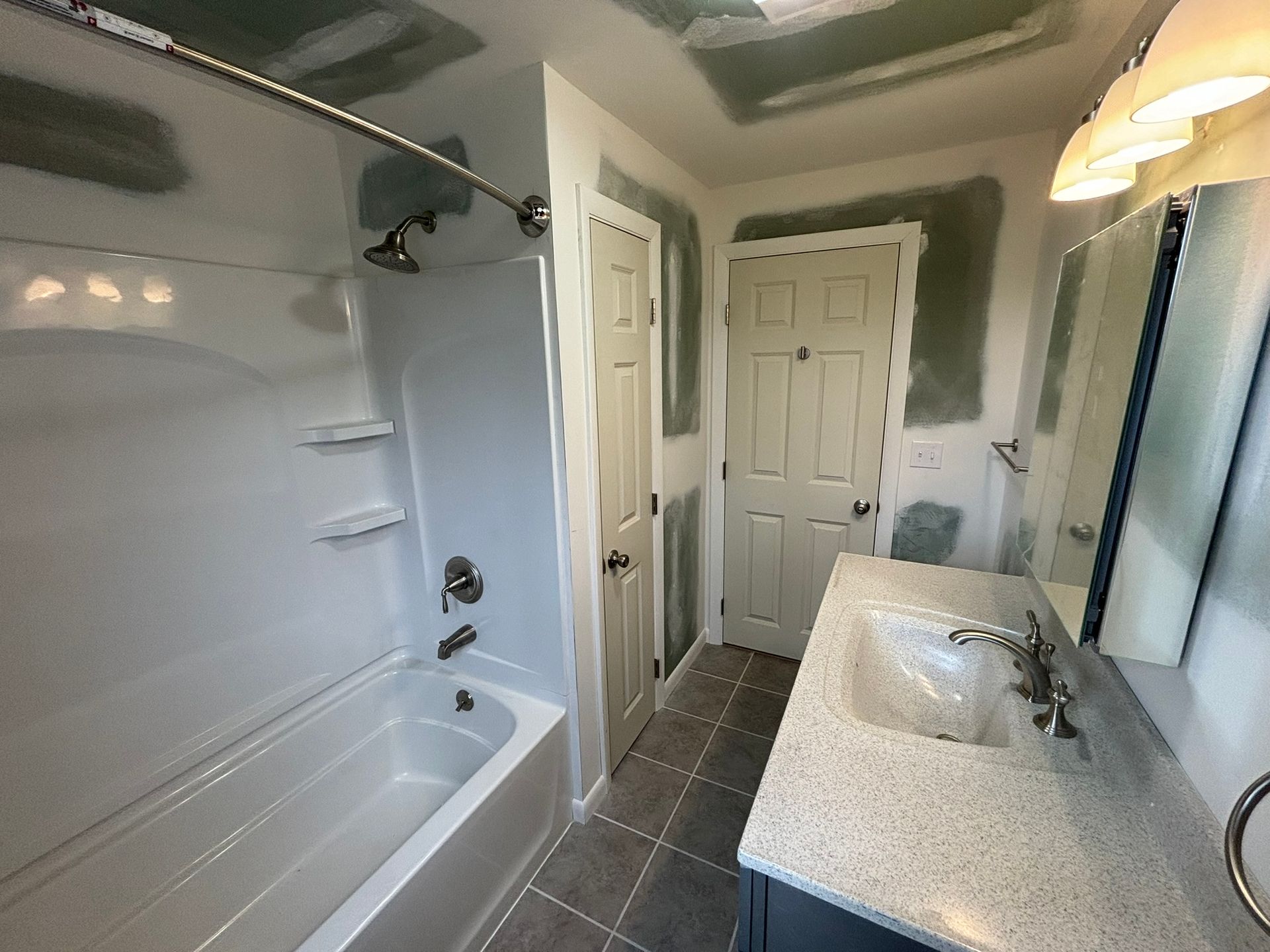 Small bathroom with white tub, gray tile floor, and speckled vanity countertop under a mirror.
