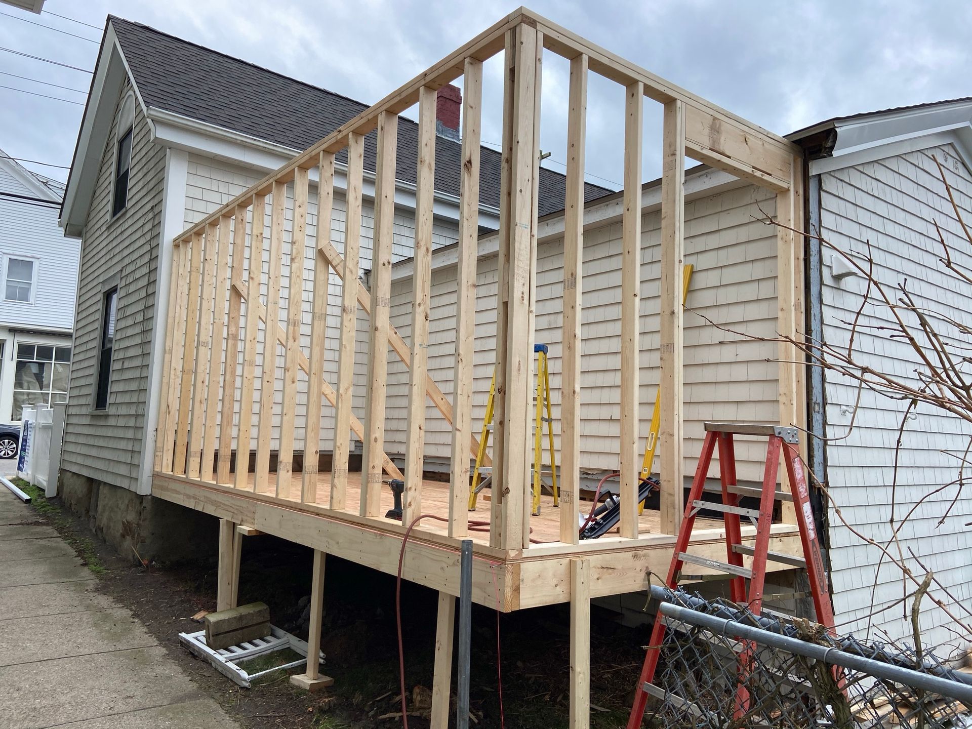 Raised wooden deck under construction beside a house, with exposed framing and a ladder nearby.