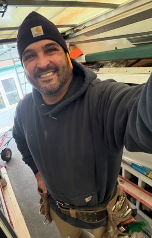 Smiling worker in a black beanie and hoodie taking a selfie inside a workshop frame