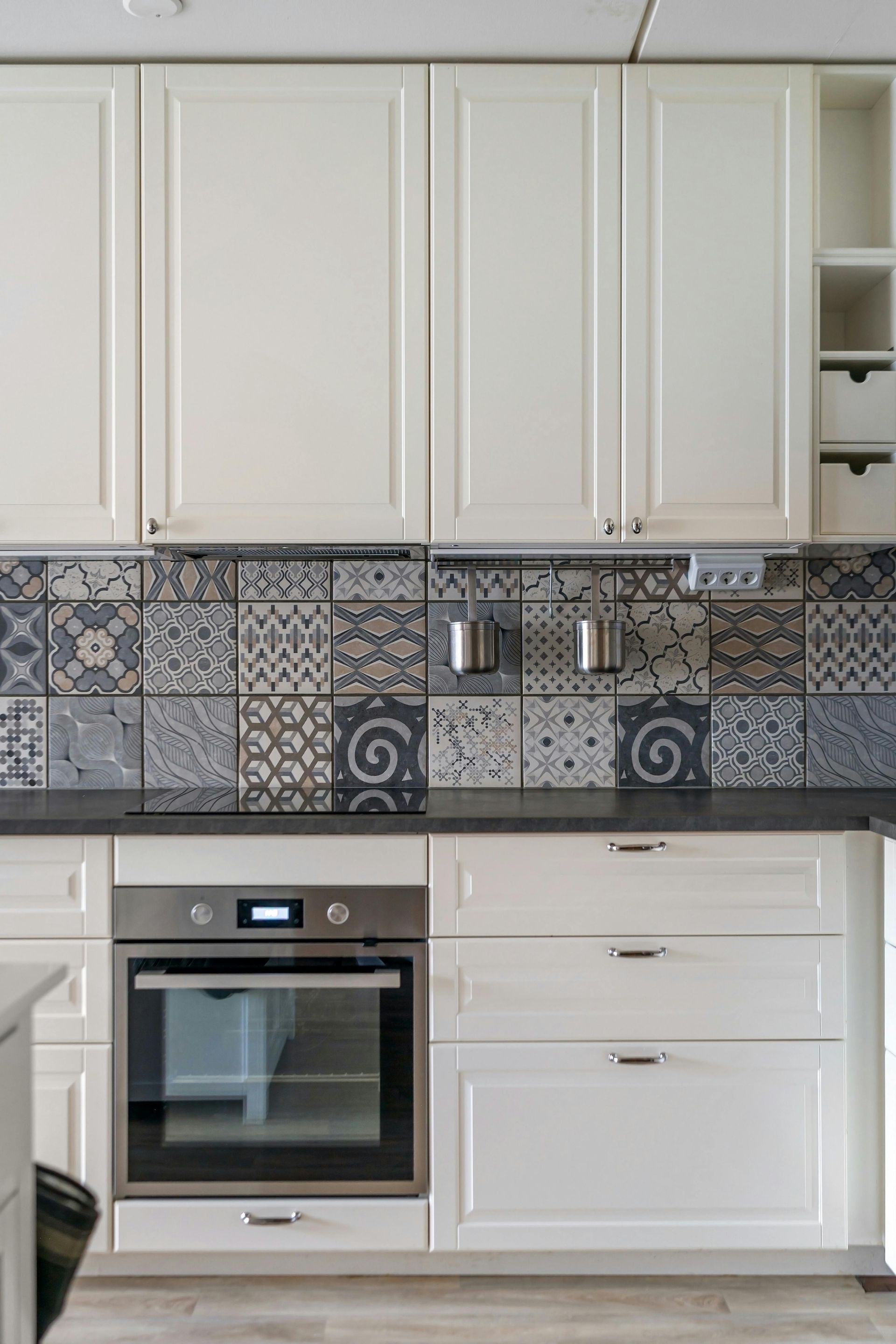Modern white kitchen cabinets with patterned blue-gray tile backsplash, built-in oven, and countertop spice rack