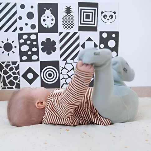 Un bebé tumbado sobre una alfombra blanca, sosteniendo un peluche de elefante, con bloques de un estampado blanco y negro detrás.