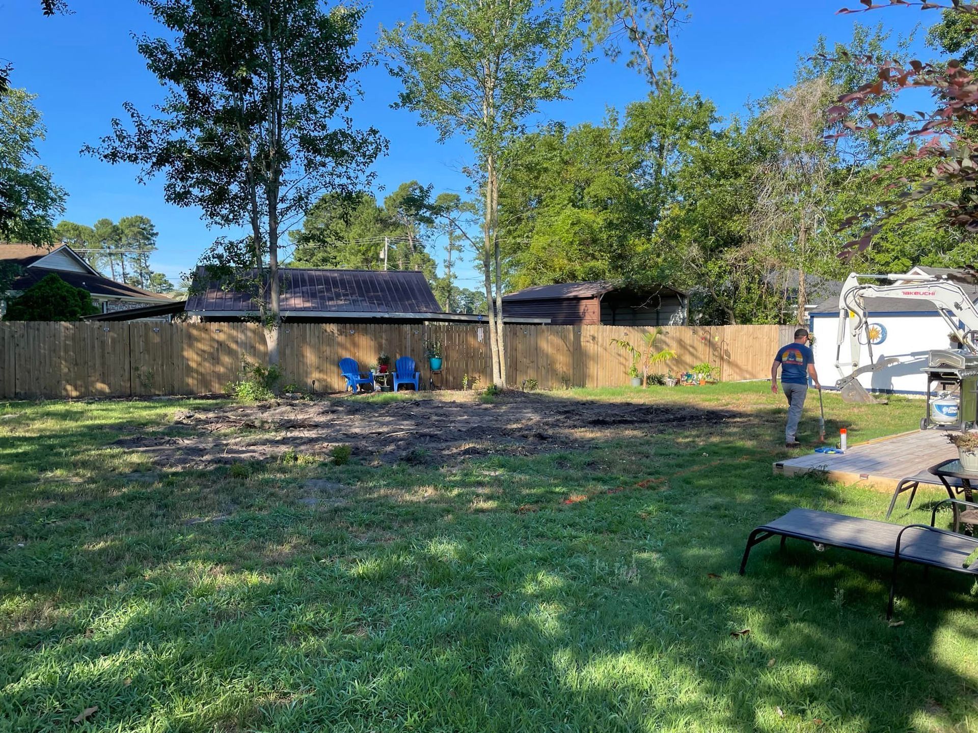 Man raking a newly cleared area in a backyard, near a wooden fence and lawn chairs. Blue sky.