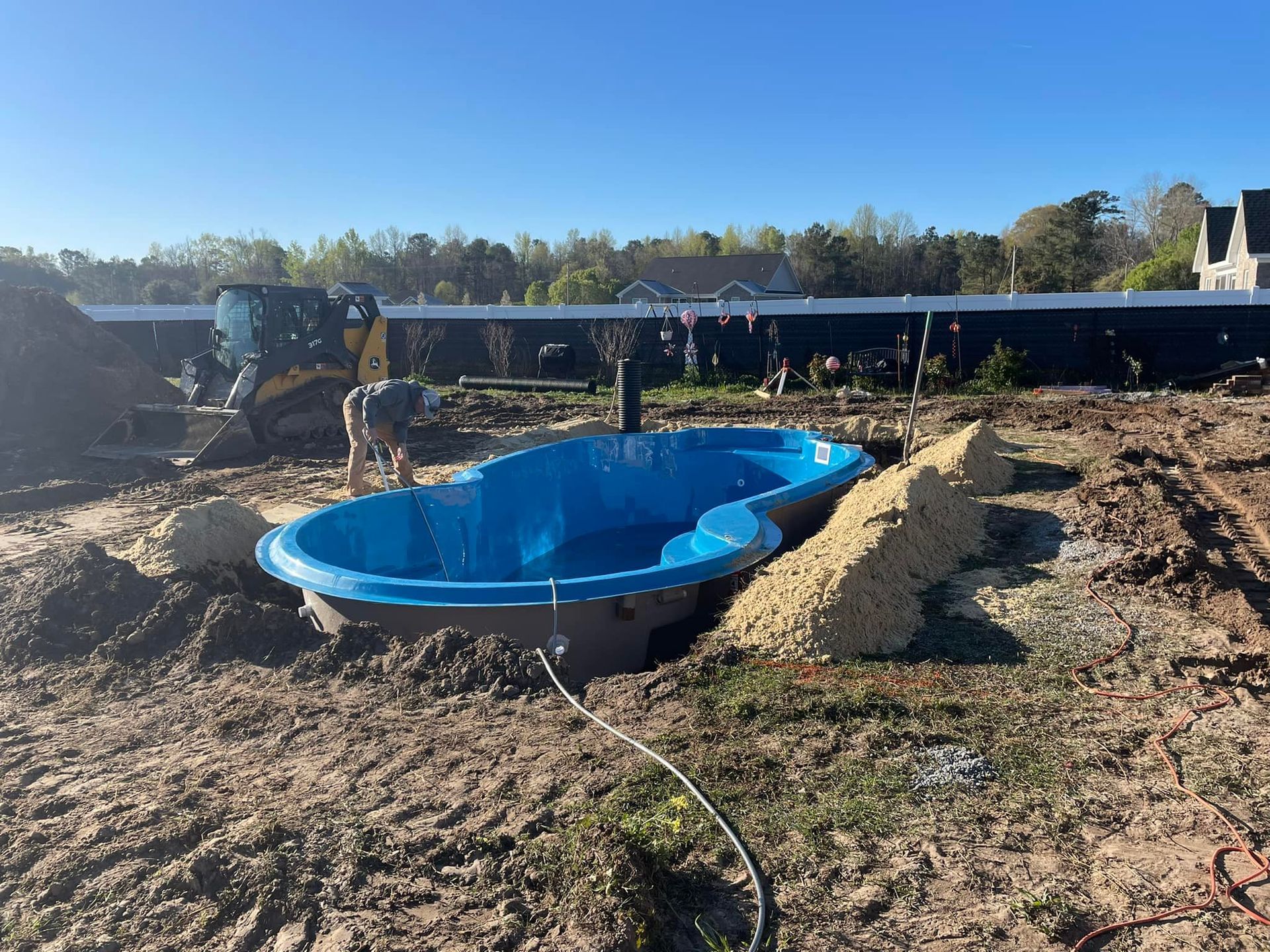 A blue, kidney-shaped pool shell sits in a dirt excavation, with a small excavator visible nearby.